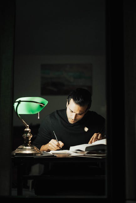Concentrated young male wearing black turtleneck taking notes and reading books while sitting at desk with papers and bankers lamp in dark home office